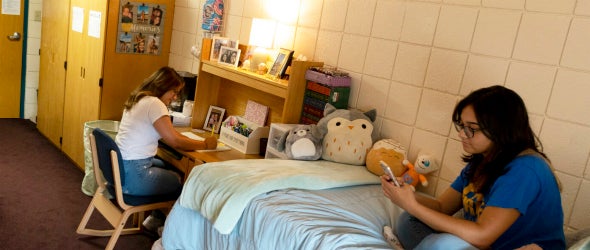 Two Students in college hall room, one seated at desk, the other on bed