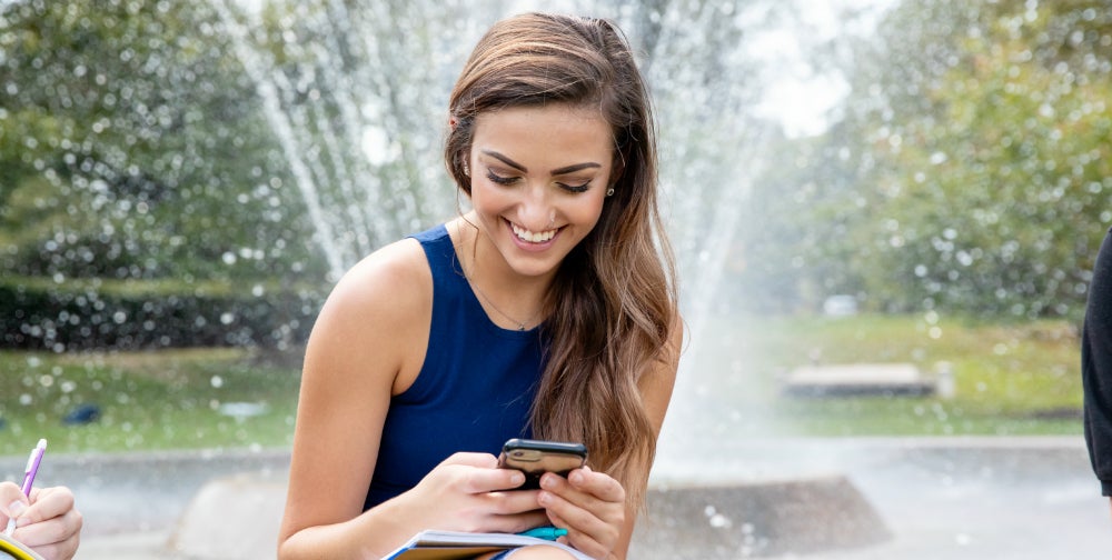 Student seated near campus fountain using smartphone.