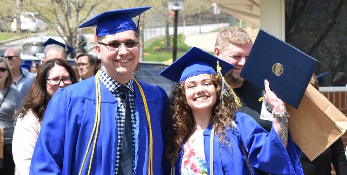 Two students in blue graduation cap and gown smiling, student on the right holding up diploma