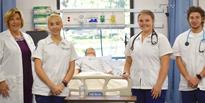 Nursing professor and three nursing students in white outfits smiling at camera. Nursing instruction equipment in the background.